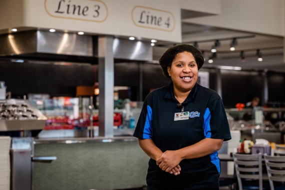 Happy woman stands with hands crossed in front of food serving line.